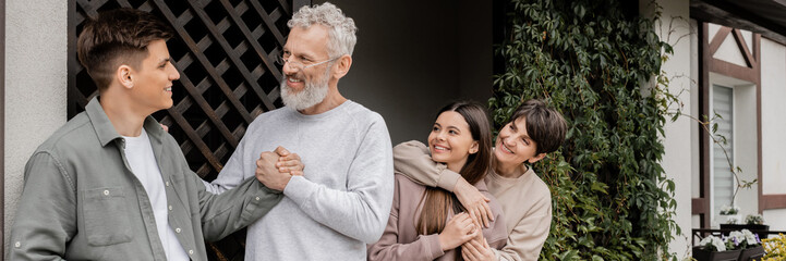 Smiling middle aged man shaking hand of young son while wife hugging daughter during parents day celebration near house at backyard, parent-child relationship concept, banner