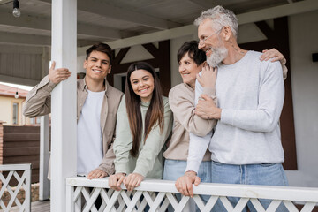 parents day, middle aged parents hugging and looking away near teenage daughter and adult son on porch of summer house, family celebration, bonding, modern parenting, translation of tattoo: harm none - obrazy, fototapety, plakaty