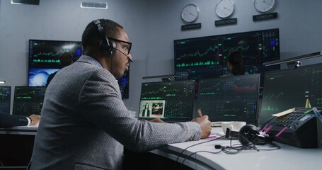 African American trader with pen in hand works at computer with displayed real-time stocks. Colleagues analyze exchange market charts on big screens at background. Concept of trading and investment.