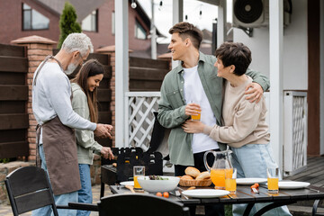 modern parenting, happy parents day, middle aged mother hugging with cheerful young adult son holding glass of orange juice, father and teenage daughter preparing food on bbq grill, summer, backyard