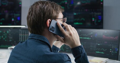 Male stock trader talks by phone, works in broker office. Computer monitors and big digital screens showing real-time stocks, exchange market charts. Investment, cryptocurrency trading and analytics.