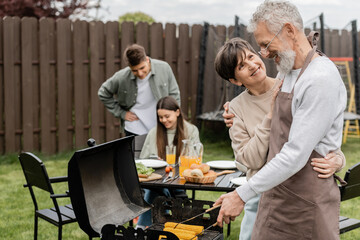 parents day, middle aged couple smiling during family bbq party, bearded tattooed man holding tongs near bbq grill, grilling corn, daughter and son, translation of tattoo: harm none do what you will - obrazy, fototapety, plakaty