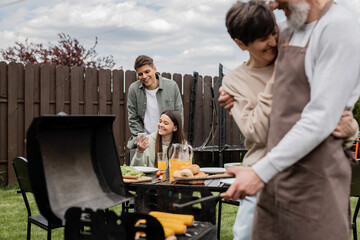 happy teenage girl showing something on smartphone to young adult brother, digital age, father preparing food on bbq grill, barbecue party, parents day cerebration, backyard, candid