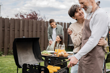 parents day, middle aged couple hugging during family bbq party, bearded tattooed man holding tongs near bbq grill, grilling corn, daughter and son, translation of tattoo: harm none do what you will - obrazy, fototapety, plakaty