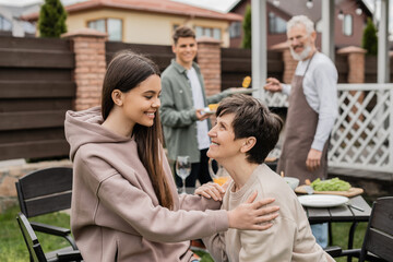 cheerful teenage girl talking to middle aged mother during family grill party, bonding, father and son preparing food on bbq grill, celebrating parents day concept, backyard of summer house