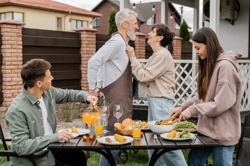 happy middle aged couple looking at each other during family bbq party, young adult son looking at cheerful parents, love, joyful teenage girl mixing salad, happy parents day, backyard, summer