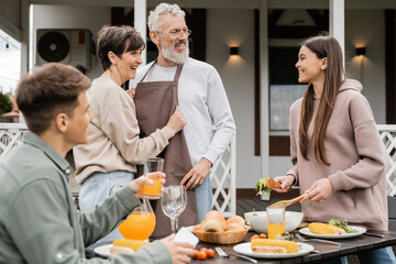 Fototapeta na wymiar celebrating parents day, happy middle aged couple looking at joyful teenage girl serving salad near adult brother with glass of orange juice, love, family grill party, summer