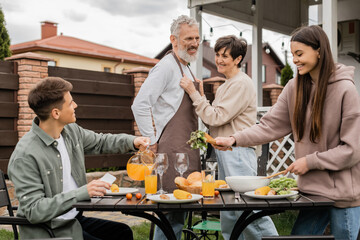 happy middle aged couple on backyard, joyful teenage girl serving salad near adult brother pouring orange juice, celebrating parents day, love, family grill party, summer