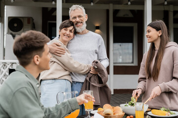 happy marriage, celebrating parents day, cheerful middle aged couple hugging near joyful teenage daughter mixing salad next to adult brother, love, family grill party, summer,  summer house backyard