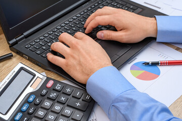 Mężczyzna pracujący przy biurku, piszący na komputerze , obok leżący kalkulator i wykres kołowy 