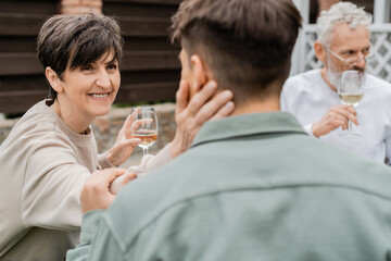 Smiling middle aged mother holding glass of wine and touching blurred adult son near husband at background during parents day celebration at backyard, family love and unity concept