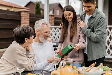 Smiling siblings giving present box to middle aged parents near summer food during barbeque party and parents day celebration at backyard in june, celebrating parenthood day concept
