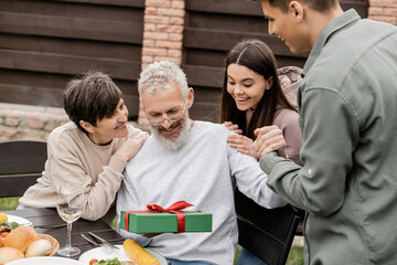 Smiling middle aged man holding gift box near children and wife during barbeque party with summer food and parents day celebration at backyard in june, celebrating parenthood day concept - Powered by Adobe