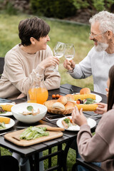 Smiling middle aged wife and husband toasting glasses of wine near summer food and teenage daughter during bbq party and parents day celebration at backyard, cherishing family bonds concept