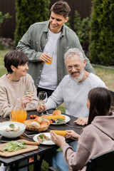 Smiling kids talking to middle aged parents near tasty summer food and parents day celebration during barbeque party at backyard in june, cherishing family bonds concept, special occasion