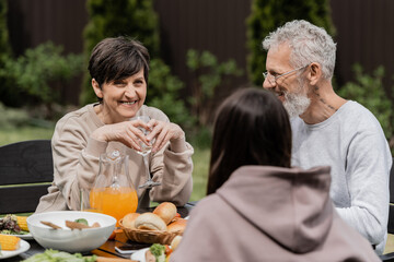 Cheerful middle aged woman holding wine glass and siting near blurred daughter and husband during summer bbq party and parents day celebration at backyard, cherishing family bonds concept