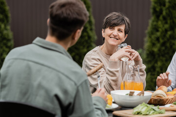 Smiling middle aged woman holding glass of wine and looking at blurred young son near husband and summer food during parents day celebration at backyard, cherishing family bonds concept