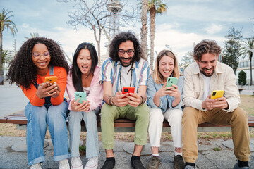 Group of best friends enjoying and smiling using their mobile phone app sitting on a bench sharing...