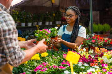 Happy black ethnicity female customer buying flowers from gardener in nursery inside greenhouse