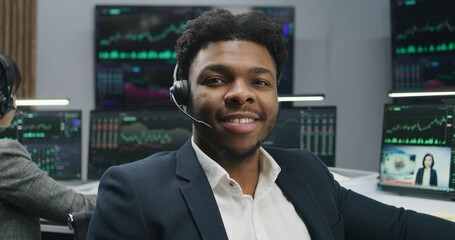Fototapeta premium African American trader in headset sits near computer with displayed real-time stocks and looks at camera. Colleagues analyze exchange market charts at background. Investment and trading. Portrait.