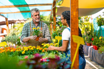 Happy female customer buying yellow flowers from a gardener in a nursery inside the greenhouse - obrazy, fototapety, plakaty