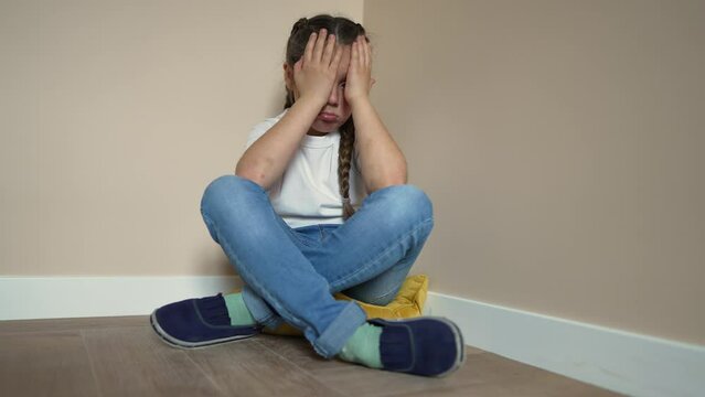School-age child is sad in corner of room. Sad, crying child offended. girl is sitting in corner on floor of room indoors. Punishment of children at home in family. Brutal upbringing at home