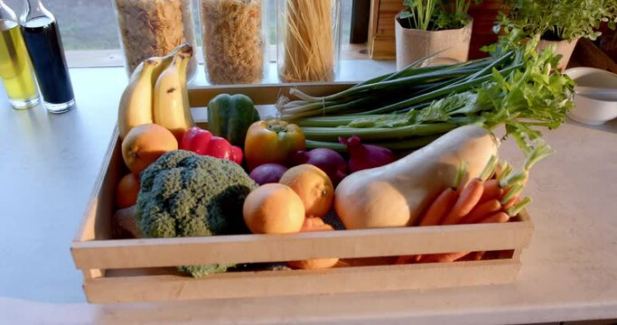 Crate of organic vegetables and storage jars of food on countertop in sunny kitchen, slow motion
