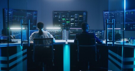 Back view of diverse IT technical specialists working on computers with database server in modern monitoring office at night. Big digital screens with real-time analysis charts and blockchain network.