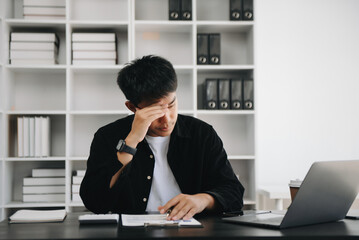 Frustrated young Asian businessman working on a laptop computer sitting at his working place in modern office