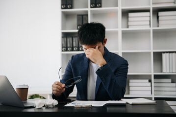 Frustrated young Asian businessman working on a laptop computer sitting at his working place in modern office