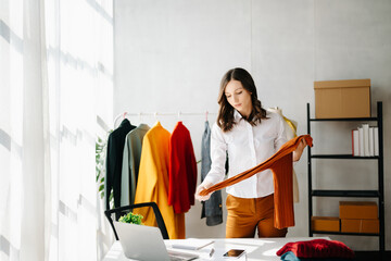 Fashion designer woman talking smart phone and using laptop with digital tablet computer in modern studio the clothes hanging on the racks .