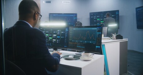 Male African American engineer types keyboard and analyzes displayed server data and real-time charts. Multi-cultural big data IT specialists work at background. Concept of cyber security. Back view.