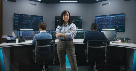 Female big data scientist stands in monitoring room and looks at camera. Software engineers work at...