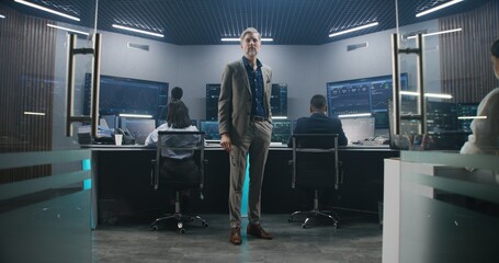 Mature big data scientist puts on glasses, stands in monitoring room and looks at camera. Software engineers work at computers at background. Multiple big screens with displayed live analysis feed.