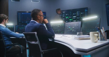 Multiethnic IT technical specialists sit in front of computers with data server in monitoring office. Software engineer works with live analysis charts and blockchain network on big digital screens.