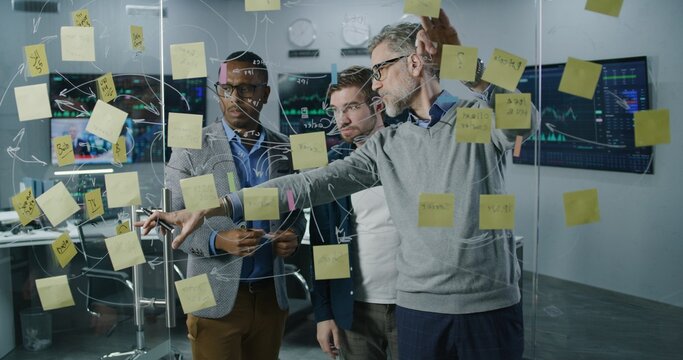 Team of diverse stock traders draw on glass wall, develop and analyze business investment strategy in broker agency office. Computers and big digital screens with exchange market charts on background.