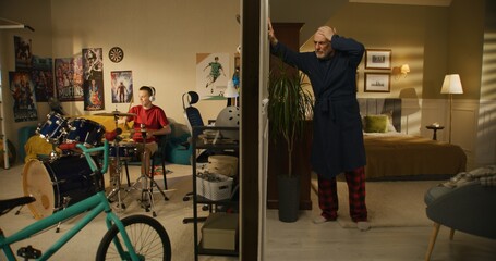 Young boy plays on drum kit at home. Elderly man irritated by noisy neighbor, screams, knocks on...