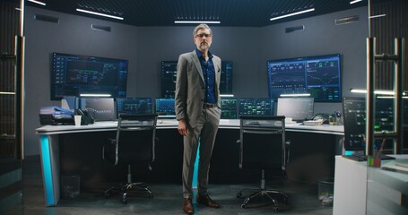 Mature big data scientist in business suit puts on glasses, stands in monitoring room and looks at camera. Multiple big screens on the wall with displayed live analysis feed at background. Dolly shot.