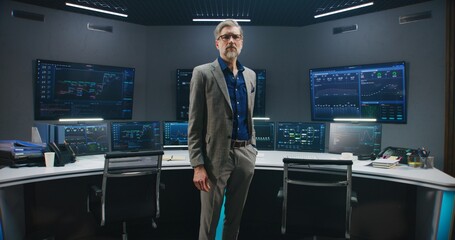 Mature big data scientist in business suit puts on glasses, stands in monitoring room and looks at camera. Multiple big screens on the wall with displayed live analysis feed at background. Dolly shot.