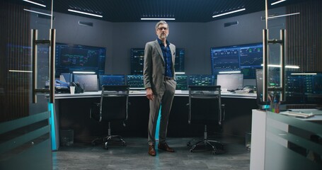 Mature big data scientist in business suit puts on glasses, stands in monitoring room and looks at camera. Multiple big screens on the wall with displayed live analysis feed at background. Dolly shot.