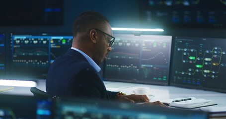 African American software engineer sits in front of computer and big digital screens with data...