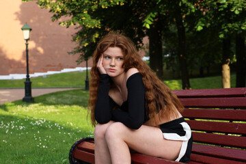 Bored girl with long red hair and freckles wearing shorts and black top sitting on a bench in...