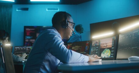 African American flight control employee in headset monitors space mission on multi-monitor computer in command center. Team clap hands after successful space rocket launch displayed on big screens. - obrazy, fototapety, plakaty