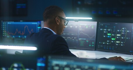 African American software engineer sits in front of computer and big digital screens with data server and blockchain network, works in modern monitoring control room with real-time analysis charts.