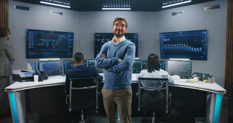 Adult IT scientist smiles, stands in monitoring room and looks at camera. Big data engineers work at computers at background. Multiple big screens with displayed real-time analysis charts. Dolly shot.