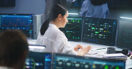 Female IT technical specialist works on computer with data server and blockchain network database in modern monitoring control room. Team of software engineers and big digital screens on background.