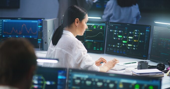 Female IT technical specialist works on computer with data server and blockchain network database in modern monitoring control room. Team of software engineers and big digital screens on background.