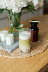 Home cozy interior. Candles, a bouquet of peonies in a glass vase, a glass of matchalate on a wooden coffee table. Spring.