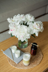 Home cozy interior. Candles, a bouquet of peonies in a glass vase, a glass of matchalate on a wooden coffee table. Spring.