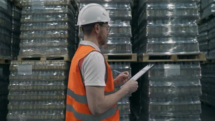 Storehouse worker counting the goods shipment at the warehouse. Worker checks the notepad to inspect the goods supply at the storehouse. Storehouse Worker walking besides the goods.